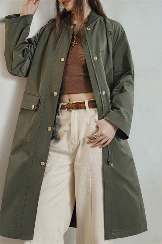 Person wearing a green trench coat over a brown sweater and white pants on a plain background
