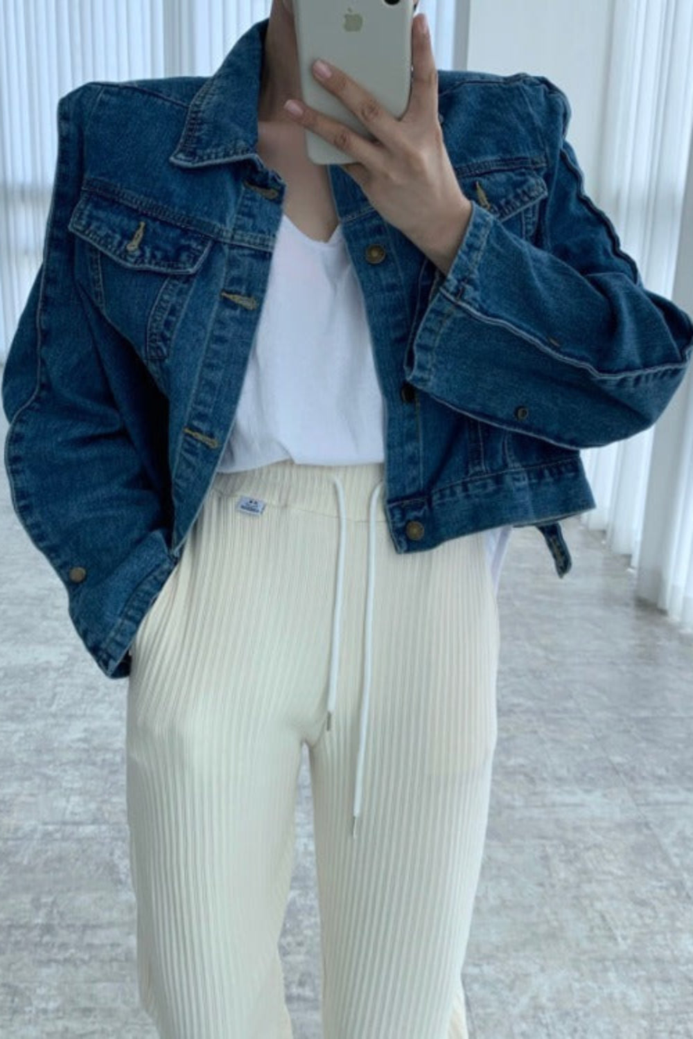 Person wearing a blue denim jacket and white pants in a minimalistic room.