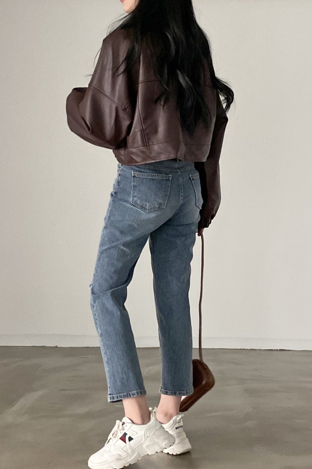 Person wearing a brown leather jacket, blue jeans, and white sneakers on a plain background