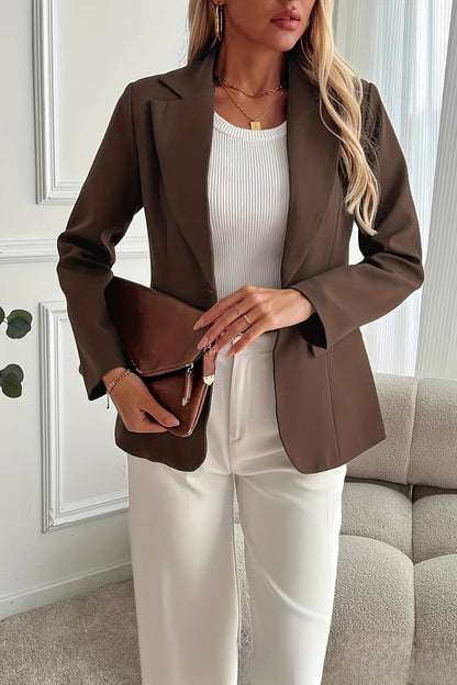 Woman in a brown blazer and white pants holding a brown clutch in a room with a gray sofa and white walls.
