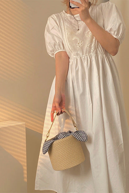 Person wearing a white dress holding a woven bag with a checkered detail against a warm-toned wall.