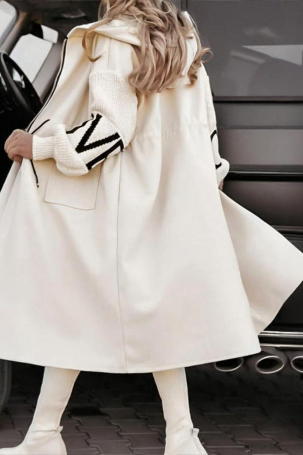 Person wearing a long cream coat with black and white striped sleeves, standing next to a car.