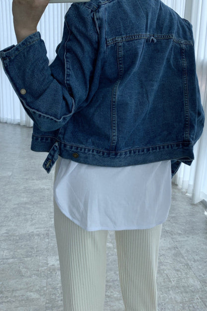 Person wearing a blue denim jacket over a white shirt and beige pants in an indoor setting.