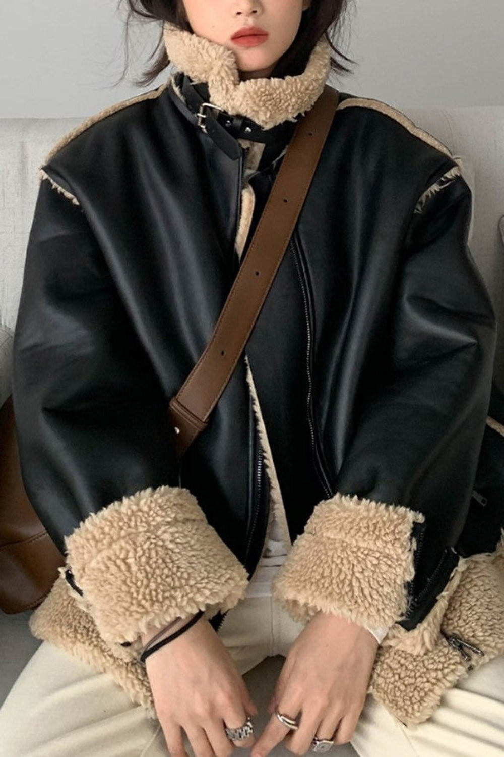 Person wearing a black leather jacket with beige fur trim sitting on a gray couch.